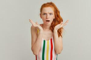 Frowning lady, discontent woman with ginger pony tail and freckles. Wearing striped colorful swimsuit. Emotion concept. Shocked with what she sees. Watching at the camera isolated over grey background photo