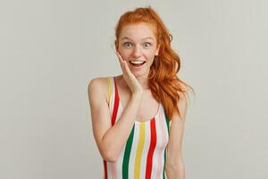 Portrait of attractive, amazed girl with ginger pony tail and freckles. Wearing striped colorful swimsuit. Touching her cheek in surprise. Watching at the camera isolated over grey background photo