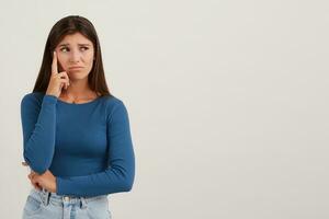 Portrait of discontent, unhappy girl with dark long hair. Wearing blue jumper and jeans. Touching her temple, thinking. Watching to the right at copy space, isolated over white background photo