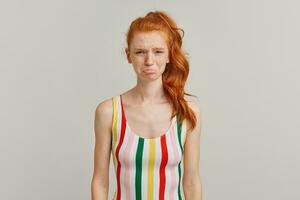 Portrait of unhappy, crying girl with ginger pony tail and freckles. Wearing striped colorful swimsuit. Emotion concept. Pouts her lip. Watching at the camera isolated over grey background photo