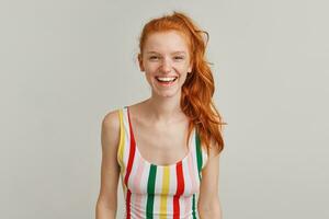 Nice looking woman, cheerful redhead girl with pony tail and freckles. Wearing striped colorful swimsuit. Emotion concept. Smiling and laughing. Watching at the camera isolated over grey background photo