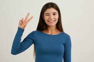 Cute looking woman, beautiful girl with dark long hair. Wearing blue jumper. Emotion concept. Shows peace sign and winks. Watching at the camera isolated over white background photo