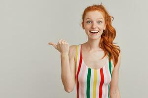 Portrait of amazed girl with ginger pony tail and freckles. Wearing striped colorful swimsuit. Watching at the camera and pointing with thumb to the left at copy space, isolated over grey background photo