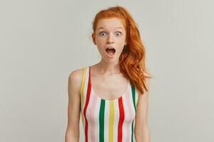 Portrait of shocked, amazed girl with ginger pony tail and freckles. Wearing striped colorful swimsuit. Emotion concept. Opens mouth in surprise. Watching at the camera isolated over grey background photo