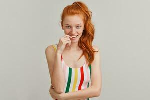 Nice looking woman, flirty redhead girl with pony tail and freckles. Wearing striped colorful swimsuit. Emotion concept. Bites finger. Watching tempting at the camera isolated over grey background photo