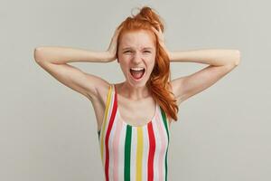 Screaming lady, stressed woman with ginger pony tail and freckles. Wearing striped colorful swimsuit. Emotion concept. Touching her head and shout. Watching at the camera isolated over grey background photo