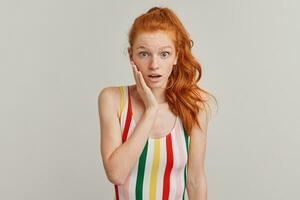 Shocked looking woman, beautiful redhead girl with pony tail and freckles. Wearing striped colorful swimsuit. Emotion concept. Touching cheek. Watching at the camera isolated over grey background photo