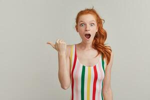 Shocked, beautiful redhead girl with pony tail and freckles. Wearing striped colorful swimsuit. Watching at the camera and pointing with thumb to the left at copy space, isolated over grey background photo
