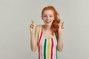 Positive woman, beautiful redhead girl with pony tail and freckles. Wearing striped colorful swimsuit. Keeps fingers crossed, make a wish. Watching at the camera isolated over grey background photo