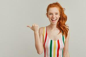 Cheerful, pretty woman with ginger pony tail and freckles. Wearing striped colorful swimsuit. Watching at the camera and pointing with thumb to the left at copy space, isolated over grey background photo