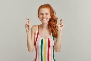 Portrait of attractive, squinting girl with ginger pony tail and freckles. Wearing striped colorful swimsuit. Keeps fingers crossed, make a wish. Stand with closed eyes isolated over grey background photo