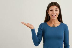 Positive looking woman, beautiful girl with dark long hair. Wearing blue jumper. Emotion concept. Watching at the camera and points palm to the left at copy space, isolated over white background photo