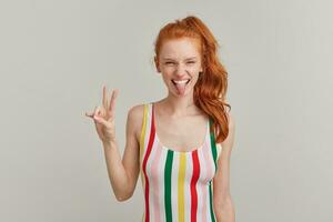 Portrait of attractive, playful girl with ginger pony tail and freckles. Wearing striped colorful swimsuit. Shows peace sign and tongue, squints. Watching at the camera isolated over grey background photo