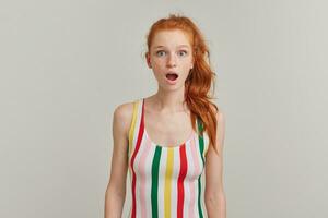 Shocked woman, beautiful redhead girl with pony tail and freckles. Wearing striped colorful swimsuit. Emotion concept. Opens mouth in amazement. Watching at the camera isolated over grey background photo