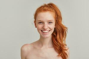 Portrait of attractive, cheerful girl with ginger pony tail and freckles. Has no make up and stand shirtless. Emotion concept. Watching at the camera and smile. Close up isolated over grey background photo