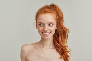 Cute looking woman, beautiful redhead girl with pony tail and freckles. Has no make up and stand shirtless. Emotion concept. Watching to the left at copy space. Close up isolated over grey background photo