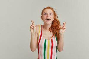 Dreaming lady, pretty woman with ginger pony tail and freckles. Wearing striped colorful swimsuit. Keeps fingers crossed, make a wish. Watching up at copy space, isolated over grey background photo