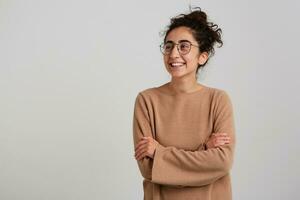 Portrait of attractive, adult girl with dark curly hair bun. Wearing beige jumper and glasses. Emotion concept. Keeps arms crossed. Watching to the left at copy space, isolated over white background photo