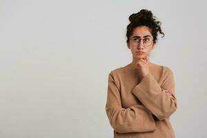 Thinking woman, beautiful girl with dark curly hair bun. Wearing beige jumper and glasses. Emotion concept. Touching chin and watching to the left at copy space, isolated over white background photo