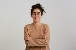 Cheerful lady, pretty woman with dark curly hair bun. Wearing beige jumper and glasses. Emotion concept. Keeps arms crossed on a chest and smile. Watching at the camera isolated over white background photo