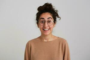 Excited lady, surprised woman with dark curly hair bun. Wearing beige jumper and glasses. Emotion concept. Happy with what she sees. Watching at the camera isolated over white background photo