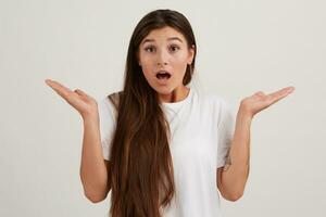 Young lady, shocked woman with dark long hair. Wearing white t-shirt. Has tattoo. Emotion concept. Shrugs her hands, unsure what to do. Watching at the camera isolated over white background photo