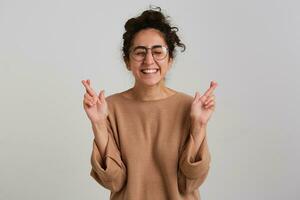 Young lady, pretty woman with dark curly hair bun. Wearing beige jumper and glasses. Emotion concept. Keeps fingers crossed and eyes closed, make a wish. Stand isolated over white background photo