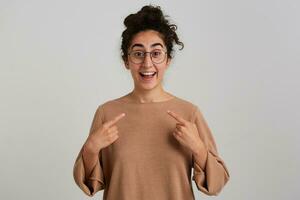 retrato de feliz, positivo niña con oscuro Rizado pelo bollo. vistiendo beige saltador y lentes. emoción concepto. señalando con dedos a sí misma. acecho a el cámara aislado terminado blanco antecedentes foto