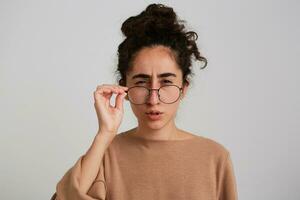 Suspicious looking woman, serious girl with dark curly hair bun. Wearing beige jumper and glasses. Emotion concept. Touching glasses and watching over them at the camera isolated over white background photo