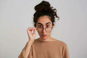 Shocked lady, pretty woman with dark curly hair bun. Wearing beige jumper. Touching glasses and gaze at you. Emotion concept. Watching at the camera, isolated close up over white background photo