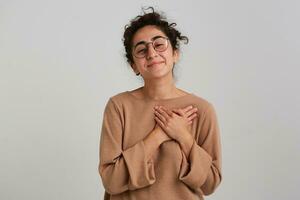 Portrait of attractive, lovely girl with dark curly hair bun. Wearing beige jumper and glasses. Emotion concept. Puts hands on a heart. Watching at the camera isolated over white background photo