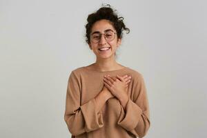 bonito mirando mujer, hermosa niña con oscuro Rizado pelo bollo. vistiendo beige saltador y lentes. emoción concepto. pone manos en un corazón y sonrisa. acecho a el cámara aislado terminado blanco antecedentes foto