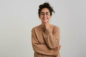 Happy lady, positive woman with dark curly hair bun. Wearing beige jumper and glasses. Emotion concept. Touching her chin and smile. Watching at the camera isolated over white background photo