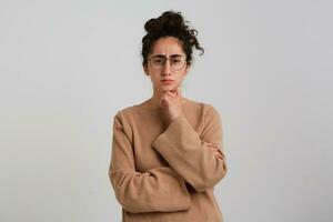 Portrait of serious, adult girl with dark curly hair bun. Wearing beige jumper and glasses. Emotion concept. Thoughtfully touching chin. Watching at the camera isolated over white background photo