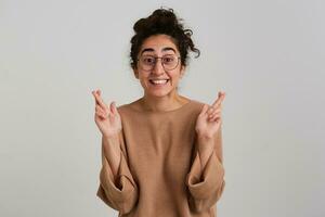 Portrait of happy, excited girl with dark curly hair bun. Wearing beige jumper and glasses. Emotion concept. Keeps fingers crossed, make a wish. Watching at the camera isolated over white background photo