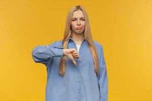 Frowning girl, discontent looking woman with blond long hair. Wearing blue shirt. People and emotion concept. Showing thumb down, disapproval. Watching at the camera, isolated over orange background photo