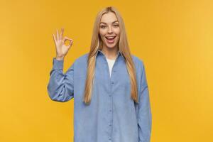 Nice looking woman, beautiful girl with long blond hair. Wearing blue shirt. People and emotion concept. Showing okay, all good sign. Watching at the camera, isolated over orange background photo