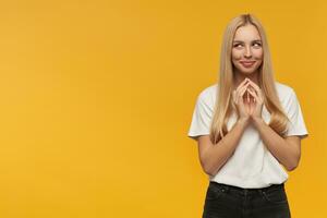 beautiful girl with long blond hair. Wearing white t-shirt and black jeans. keeps her palm together, think about some ideas, Watching to the left at copy space, isolated over orange background photo