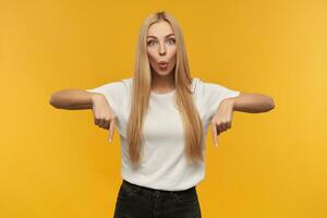 Teenage girl, happy looking woman with blond long hair. Wearing white t-shirt and black jeans. People and emotion concept. Watching at the camera and pointing down at copy space photo