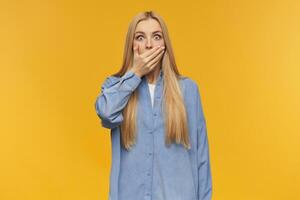 Portrait of shocked, adult girl with blond long hair. Wearing blue shirt. People and emotion concept. Close her mouth with palm, can't talk. Watching at the camera, isolated over orange background photo