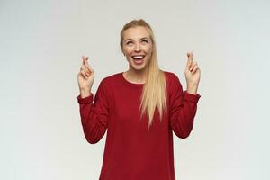 Portrait of attractive, adult girl with blond long hair. Wearing red sweater. People and emotion concept. Watching up at copy space, isolated over white wall, keeps her fingers crossed and praying photo