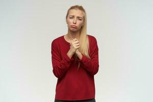 Teenage girl, unhappy looking woman with blond long hair. Wearing red sweater. People and emotion concept. Watching at the camera, isolated over white background, curving her lips and feels sad photo