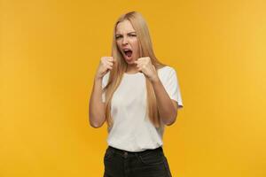 ready to fight, happy looking woman with blond long hair. Wearing white t-shirt and black jeans. People and emotion concept. Watching at the camera, isolated over orange background photo