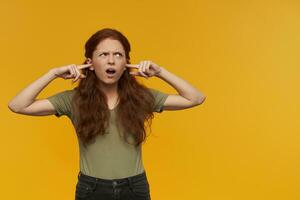 Irritated looking girl, frowning redhead woman with long hair. Wearing green t-shirt. Emotion concept. Close her ears with fingers. Watching to the right at copy space, isolated over orange background photo