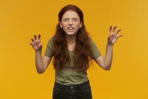 Portrait of angry, savage lady with long ginger hair. Wearing green t-shirt. Emotion concept. Pretend she has claws. Roaring like a beast. Watching at the camera, isolated over orange background photo