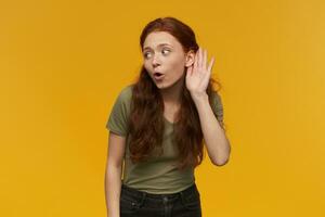 Portrait of curious, beautiful lady with long ginger hair. Wearing green t-shirt. People and emotion concept. Keeps palm next to ear, eavesdrop conversation. Stand isolated over orange background photo