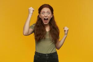 Happy looking girl, excited redhead woman with long hair. Wearing green t-shirt. People and emotion concept. Raises fists and celebrate. Watching at the camera, isolated over orange background photo