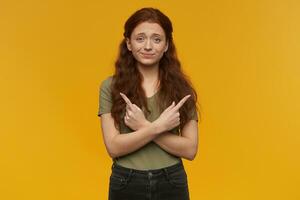 Cute, positive woman with long ginger hair. Wearing green t-shirt. People and emotion concept. Pointing uncertainly both sides at copy space. Watching at the camera, isolated over orange background photo