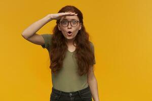 Portrait of excited, surprised lady with long ginger hair. Wearing green t-shirt and eyewear. Gaze into distance with palm over her eyes. Watching at the camera, isolated over orange background photo