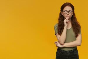 Portrait of thinking, dreaming lady with long ginger hair. Wearing green t-shirt and eyewear. Emotion concept. Touching her chin and watching to the left at copy space, isolated over orange background photo